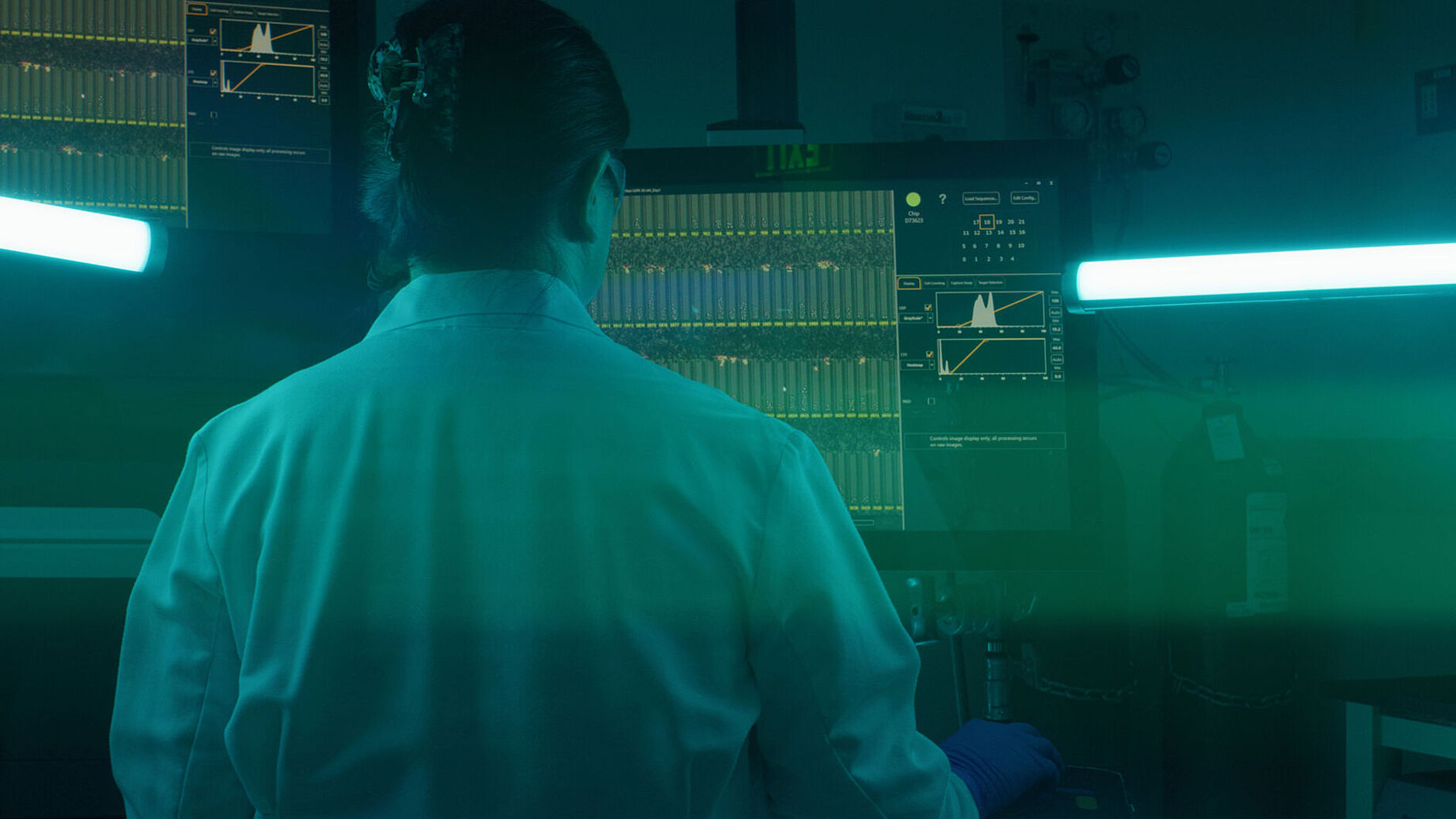 Eine Wissenschaftlerin im Laborkittel betrachtet Daten auf einem Bildschirm in einem blaugrün beleuchteten Labor.