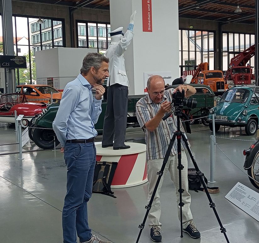 Fotoshooting im Verkehrszentrum: Zwei Männer betrachten Aufnahmen im Display einer Fotokamera umgeben von Oltimern