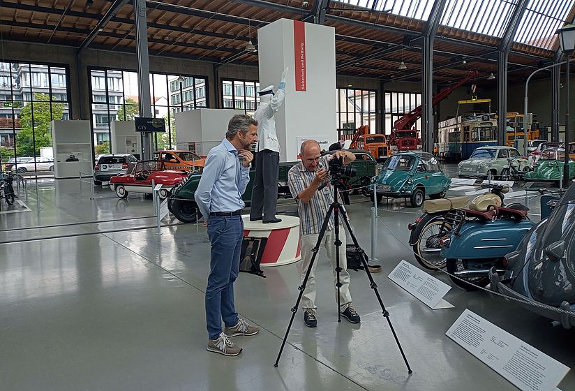Fotoshooting im Verkehrszentrum: Zwei Männer betrachten Aufnahmen im Display einer Fotokamera umgeben von Oltimern