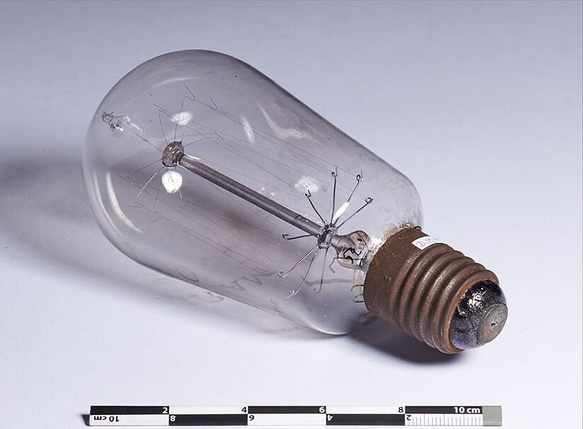 Fotografie einer alten Glühbirne, Zollstab an unterem Bildrand. Glaskörper ist birnenförmig, die Glühdrähte sind an acht Armen innerhalb aufgehängt. 