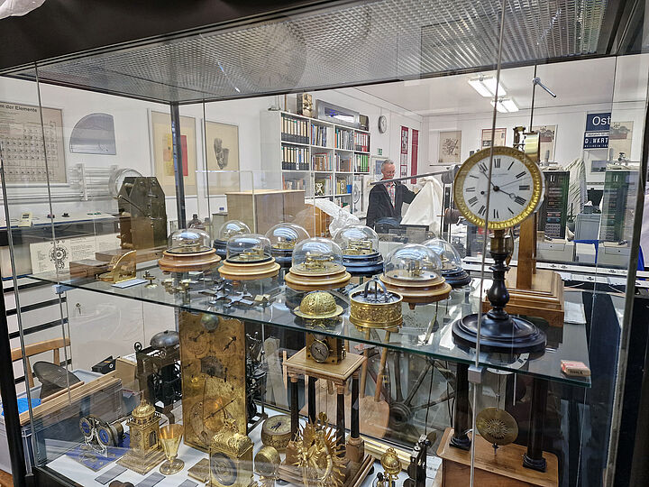 Blick durch Glasvitrine mit verschiedenen Chronometern in mit Objekten und Akten gefüllten Raum (Restaurierungswerkstatt). Person (rechts, zentral) hebt Stoffabdeckung auf.