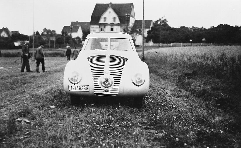 Schwarz-weiß Fotografie zeigt den BMW Wendler von vorne. Es steht auf einem Feld. Im Hintergrund befinden sich Häuser.